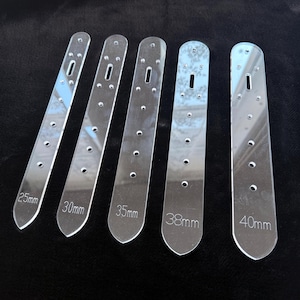 May include: Five clear acrylic belt blanks, labelled with measurements from 25mm to 40mm. Each blank has a series of small holes and a slot for a buckle. The blanks are designed for crafting belts. The measurements are in millimetres.