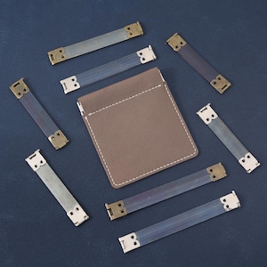 Könnte beinhalten: Eine Sammlung von Lederriemen mit Metallbeschlägen und einem braunen Lederkartenhalter. Die Riemen sind in verschiedenen Grau- und Blautönen gehalten, mit Messing- und silberfarbenen Metallakzenten. Der Kartenhalter ist braun mit weißen Nähten.