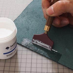 K&ouml;nnte beinhalten: Nahaufnahme eines Bastelprojekts. Ein bordeauxfarbenes Werkzeug mit Holzgriff tr&auml;gt wei&szlig;en Klebstoff auf ein St&uuml;ck teal-farbenes Leder auf. Ein wei&szlig;er Beh&auml;lter mit Klebstoff ist sichtbar, mit dem Text "ECOSTICK".