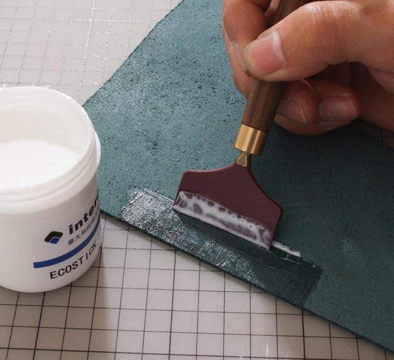 K&ouml;nnte beinhalten: Nahaufnahme eines Bastelprojekts. Ein bordeauxfarbenes Werkzeug mit Holzgriff tr&auml;gt wei&szlig;en Klebstoff auf ein St&uuml;ck teal-farbenes Leder auf. Ein wei&szlig;er Beh&auml;lter mit Klebstoff ist sichtbar, mit dem Text "ECOSTICK".