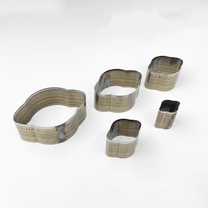 May include: Five metal leather craft tools with curved edges, arranged on a white surface. The tools have a rectangular shape with rounded corners and are labeled with text. The tools are for leather working.