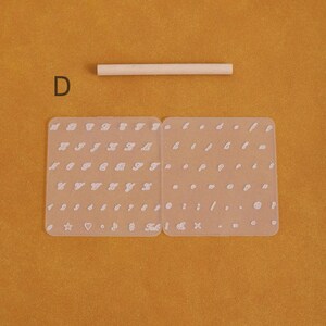 May include: A set of clear, flexible alphabet and number stamps with a wooden dowel on a mustard-colored surface. The stamps include uppercase and lowercase letters, numbers, and symbols. The letter "D" is visible in the upper left corner.