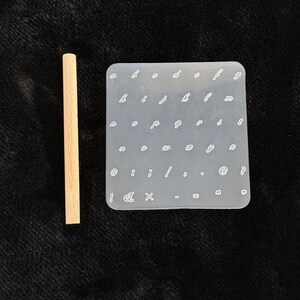 May include: A light-colored wooden dowel next to a clear, rectangular silicone mold. The mold contains various lowercase letters, numbers, and symbols in a white font. The items are on a dark surface.