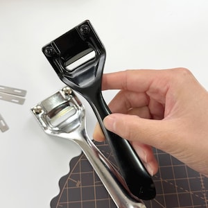 May include: Two metal blade holders, one black and one silver, are displayed. The black holder is held by a hand. The blade holders have a rectangular opening and are designed for cutting. Several spare blades are visible.