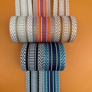 Puede incluir: Una colección de cintas tejidas en varios colores, incluyendo beige, naranja, marrón, negro y azul. Las cintas están enrolladas, mostrando sus intrincados patrones y texturas. El fondo es de color naranja sólido.