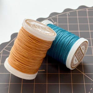 May include: Two spools of thread. One is a warm, light brown color, and the other is a teal blue. The spools have white tops with printed labels. The thread is tightly wound, and the spools are positioned at an angle against a grid background.