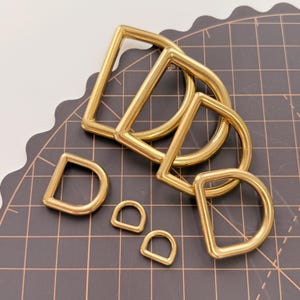 Puede incluir: Una colección de anillas en forma de D de color dorado en varios tamaños. Las anillas en D son de metal y tienen un acabado brillante. Las anillas están dispuestas sobre una esterilla de corte negra con un patrón de cuadrícula. Medidas en centímetros.