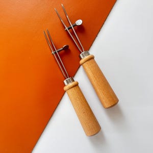 May include: Two leathercraft tools with wooden handles and metal prongs are displayed against a background of orange leather and white. The tools have a screw mechanism for adjusting the prong width, and are designed for leatherworking.
