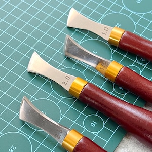 Könnte beinhalten: Vier Lederhandwerkzeuge mit Holzgriffen und Metallklingen. Die Klingen haben gebogene Kanten und sind mit Zahlen zur Größenangabe versehen. Die Werkzeuge sind diagonal auf einer grünen Schneidematte angeordnet.