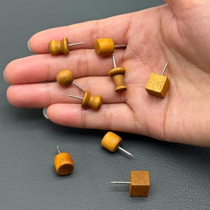 May include: A selection of small, wooden push pins in different shapes, such as cylinders, spheres, and cubes. Each pin has a silver metal needle. The pins are a light brown colour and appear to be handmade.