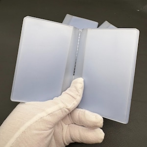 May include: A clear, rectangular plastic card holder with a hinged spine and two transparent interior sleeves. The object is constructed from a smooth, semi-translucent material and features a simple, functional design for organizing cards.