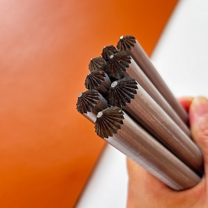 Puede incluir: Primer plano de una mano sosteniendo un grupo de herramientas de artesanía en cuero de metal. Las herramientas tienen un cuerpo cilíndrico con un extremo decorativo en forma de abanico. Un trozo de cuero naranja está en el fondo.