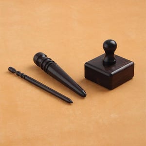 Può includere: Un set di tre utensili in legno marrone scuro, tra cui un lesina conica, uno strumento cilindrico con un'estremità arrotondata e un timbro quadrato con un manico arrotondato. Gli utensili sono disposti su una superficie marrone chiaro.