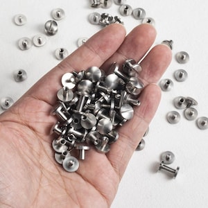 Op de afbeelding: Een verzameling kleine, zilverkleurige klinknagels met platte, ronde koppen en korte pennen. Sommige klinknagels worden in een hand gehouden, andere zijn verspreid over een wit oppervlak. De klinknagels lijken van metaal te zijn en worden waarschijnlijk gebruikt voor bevestiging.