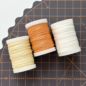 May include: Three spools of thread in shades of yellow, orange, and white. The thread is tightly wound on white plastic spools. The spools are arranged on a dark brown cutting mat with grid lines and measurement markings.