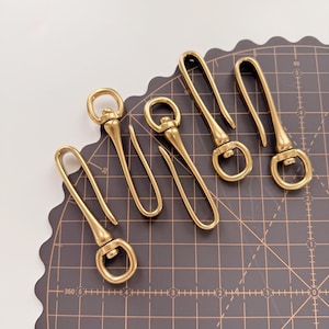 May include: A collection of six brass-colored keychains with a hook and ring design. The keychains are arranged on a dark gray cutting mat with grid lines. The keychains have a polished finish and a sturdy appearance.