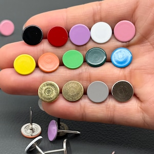 May include: A collection of colorful round push pins in various colors, including black, red, purple, white, pink, yellow, orange, green, teal, and blue. Some pins have a metallic finish. The pins are for crafts and office use.