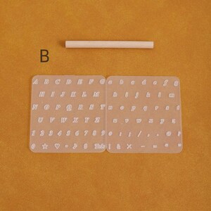 May include: A clear, rectangular alphabet and number stencil with uppercase and lowercase letters, numbers, and symbols. A small wooden dowel is above the stencil. The letter "B" is in the upper left corner.