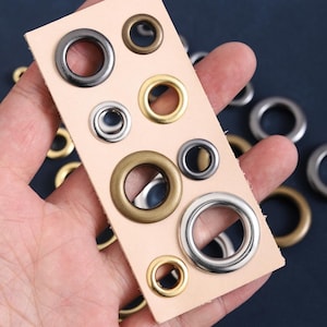 May include: Assortment of metal eyelets in various sizes and finishes, including silver, gold, and bronze. The eyelets are displayed on a piece of beige leather, showcasing their different colors and designs. Ideal for crafting and leatherwork projects.