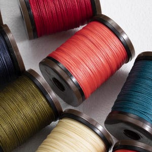 Könnte beinhalten: Eine Sammlung bunter Garnrollen. Die Rollen sind in verschiedenen Farben erhältlich, darunter Rot, Orange, Grün, Blau und Creme. Jede Rolle hat eine dunkelbraune Basis und Oberseite. Der Faden ist fest um die Rolle gewickelt.