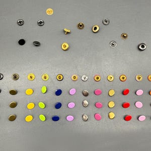 May include: A collection of metal snap fasteners in a variety of colours and finishes, including black, gold, silver, and bright hues. The snaps are arranged on a grey surface, displaying their different styles and shades.