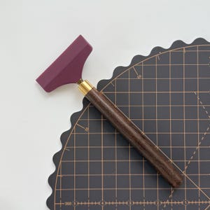 K&ouml;nnte beinhalten: Ein Lederwerkzeug mit einer weinroten Klinge, einer Messingzwinge und einem dunklen Holzgriff. Das Werkzeug dient zum Gl&auml;tten von Leder und liegt auf einer schwarzen Schneidematte mit Rasterlinien.