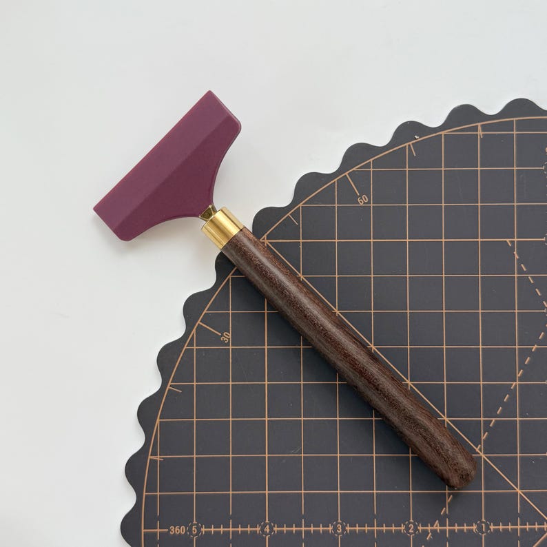 K&ouml;nnte beinhalten: Ein Lederwerkzeug mit einer weinroten Klinge, einer Messingzwinge und einem dunklen Holzgriff. Das Werkzeug dient zum Gl&auml;tten von Leder und liegt auf einer schwarzen Schneidematte mit Rasterlinien.