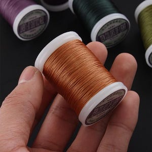 May include: A spool of copper-colored thread held in a hand, with other spools of thread in various colors in the background. The thread is tightly wound on a white plastic spool.