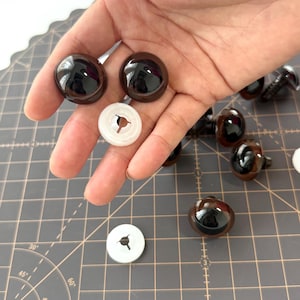 Puede incluir: Una colección de ojos de seguridad marrones y brillantes para manualidades, con arandelas de plástico blanco. Los ojos son redondos y tienen una superficie reflectante. Algunos ojos tienen un poste de tornillo en la parte posterior. Los artículos están sobre una esterilla de corte gris.