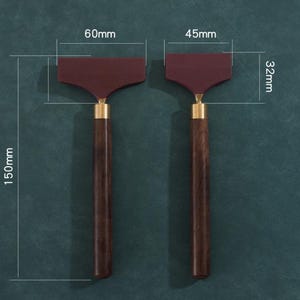 K&ouml;nnte beinhalten: Zwei dunkelbraune Holzgriff-Rakel mit weinroten Klingen und Messingakzenten. Die Rakel sind 15 cm lang, mit Klingen von 6 cm und 4,5 cm Breite und 3,2 cm H&ouml;he. Die Werkzeuge werden vor einem dunkelgr&uuml;nen Hintergrund gezeigt.