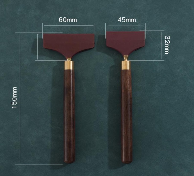 K&ouml;nnte beinhalten: Zwei dunkelbraune Holzgriff-Rakel mit weinroten Klingen und Messingakzenten. Die Rakel sind 15 cm lang, mit Klingen von 6 cm und 4,5 cm Breite und 3,2 cm H&ouml;he. Die Werkzeuge werden vor einem dunkelgr&uuml;nen Hintergrund gezeigt.