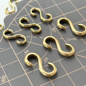 May include: Several small, shiny, brass-coloured S-hooks are scattered on a grey grid surface. The hooks are curved and have a smooth, polished finish, reflecting light. These hooks are likely used for hanging or connecting items.