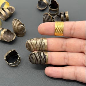 May include: A collection of antique brass thimbles in various styles. Some thimbles are open-ended, while others have a closed top. One gold-colored thimble is also present. The thimbles are designed for sewing and crafting.