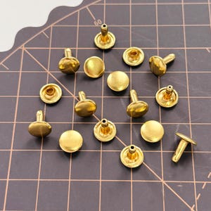 May include: A collection of small, shiny, gold-colored metal rivets. The rivets have a rounded head and a post for securing them to fabric or leather. They are scattered on a dark surface with grid lines, suggesting a crafting or sewing project.