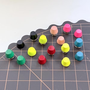 May include: A collection of colorful, cone-shaped push pins in various colors, including black, green, yellow, red, pink, and blue. The push pins are arranged on a dark gray grid surface, with a white background.