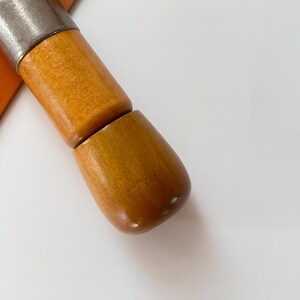 May include: Close-up of a wooden tool with a metal top. The tool has a light brown, polished wooden handle and a silver metal top. The handle is made of two connected pieces of wood. The background is white.