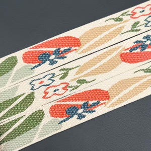 Puede incluir: Una cinta de tela tejida con una base de color blanco roto que presenta un patrón repetitivo de formas estilizadas similares a tulipanes naranjas, detalles florales azul oscuro y hojas de color verde claro y beige.