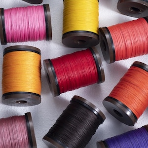 May include: A collection of colorful spools of thread, including shades of pink, yellow, red, orange, black, and purple. The thread is tightly wound on brown spools with black accents, ideal for sewing and crafting projects.