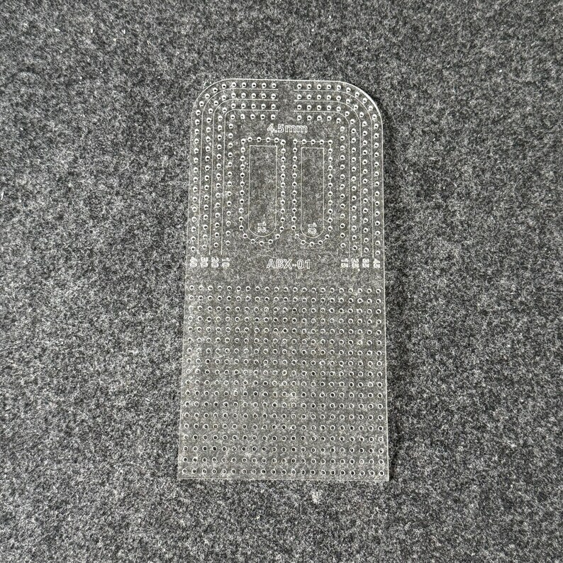 Peut inclure: Un gabarit de maroquinerie en acrylique transparent avec un bord sup&eacute;rieur arrondi et une grille de trous de couture d&eacute;coup&eacute;s avec pr&eacute;cision. L'outil porte l'inscription 4.5mm et ABX-01, ainsi que des indicateurs num&eacute;riques de guidage.