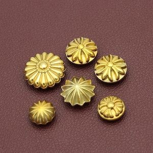 Könnte beinhalten: Sechs goldfarbene Zierknöpfe mit Blumen- und Sternmotiven. Die Knöpfe sind auf einer weinroten Oberfläche angeordnet. Die Knöpfe variieren in der Größe und haben einen metallischen Glanz.