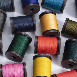 May include: A collection of colorful spools of thread, each wound around a dark brown core. The threads are in various shades, including blue, green, yellow, pink, and orange. The spools are arranged on a white surface.
