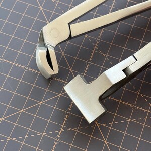 Puede incluir: Primer plano de dos herramientas de metal plateado. Una herramienta tiene una cabeza curva y redondeada con un filo, y la otra tiene una cabeza plana y rectangular. Las herramientas est&aacute;n sobre una superficie oscura con un patr&oacute;n de cuadr&iacute;cula.