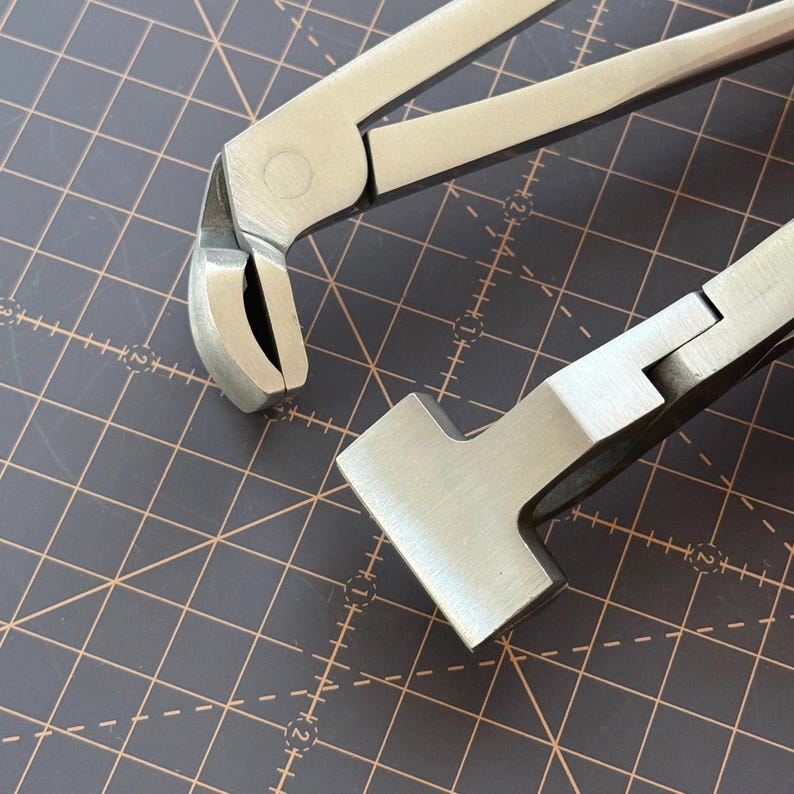 Puede incluir: Primer plano de dos herramientas de metal plateado. Una herramienta tiene una cabeza curva y redondeada con un filo, y la otra tiene una cabeza plana y rectangular. Las herramientas est&aacute;n sobre una superficie oscura con un patr&oacute;n de cuadr&iacute;cula.