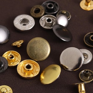 May include: Assortment of metal snap fasteners in various colors, including gold, silver, bronze, and black. The snaps are in different shapes and sizes, with some having decorative details. Ideal for sewing and crafting projects.