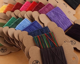 May include: A collection of colorful, 1mm diameter cord spools. The spools are arranged in rows, showcasing a variety of colors including brown, yellow, orange, red, pink, purple, blue, green, and black. Each spool is mounted on a brown paper card.