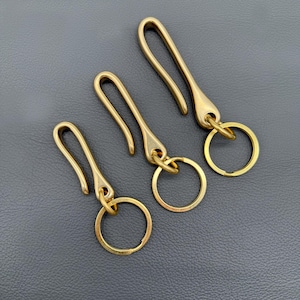 Op de afbeelding: Drie messing sleutelhangers met een haakontwerp en bevestigde sleutelringen. De sleutelhangers zijn in verschillende maten en worden gepresenteerd op een donkergrijze ondergrond. De sleutelhangers hebben een gepolijste, gouden afwerking.