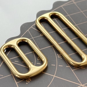 May include: Two gold-coloured rectangular slider buckles. The buckles have a rounded design with two parallel bars and a curved top. The metal has a shiny finish. One buckle is smaller than the other.