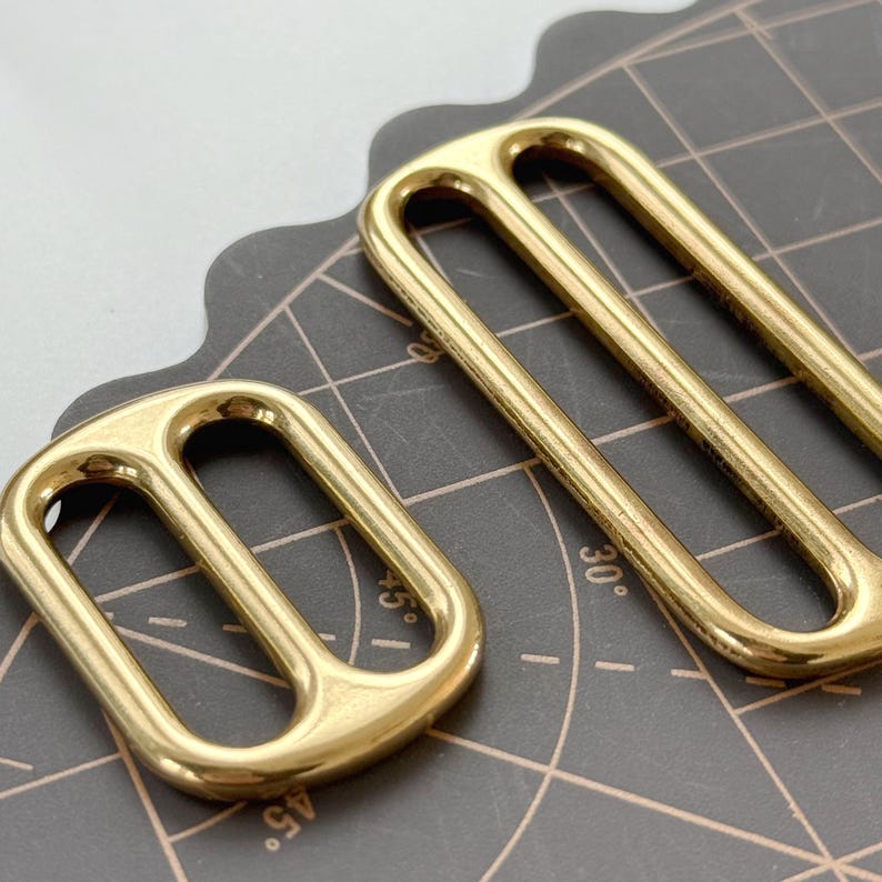 May include: Two gold-coloured rectangular slider buckles. The buckles have a rounded design with two parallel bars and a curved top. The metal has a shiny finish. One buckle is smaller than the other.