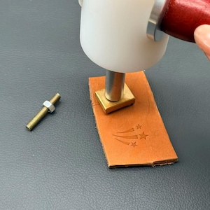 Pode incluir: Um martelo branco com cabo vermelho pressiona um carimbo de latão sobre um pedaço retangular de couro castanho. O couro apresenta um design em relevo de três estrelas com linhas. Um pequeno parafuso roscado com porca está próximo.