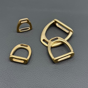 May include: Four gold-colored metal stirrups, varying in size, are arranged on a dark surface. The stirrups have a classic D-shape with a rectangular opening at the bottom and a slot at the top for attachment. The metal has a polished finish.